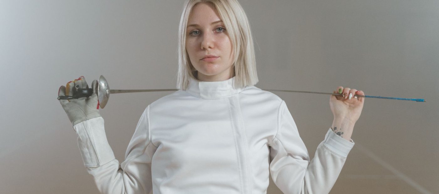 Portrait of a female fencer in white, holding an epee indoors with a determined expression.