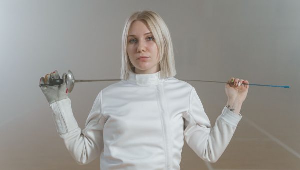 Portrait of a female fencer in white, holding an epee indoors with a determined expression.