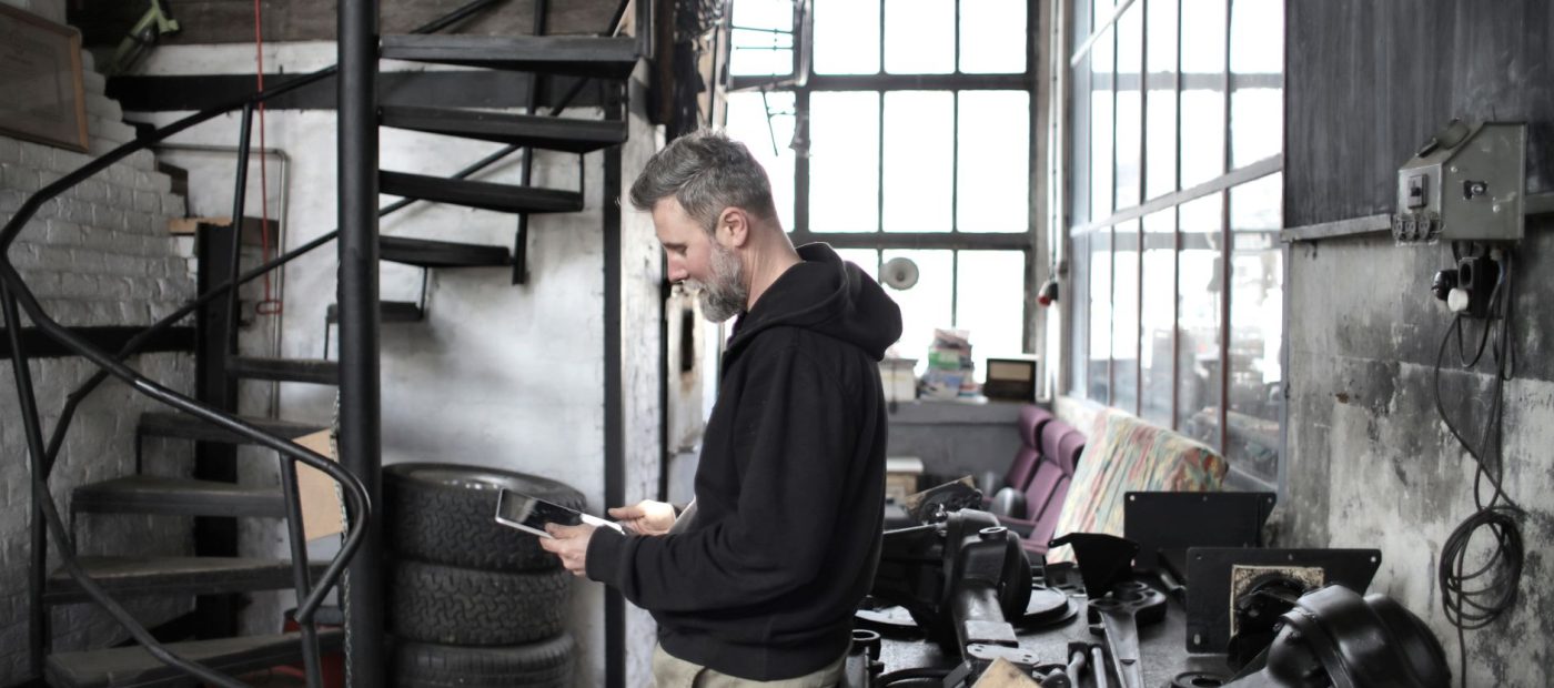 Side view of bearded adult mechanic in workwear browsing portable tablet while standing near workbench in workshop