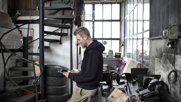 Side view of bearded adult mechanic in workwear browsing portable tablet while standing near workbench in workshop