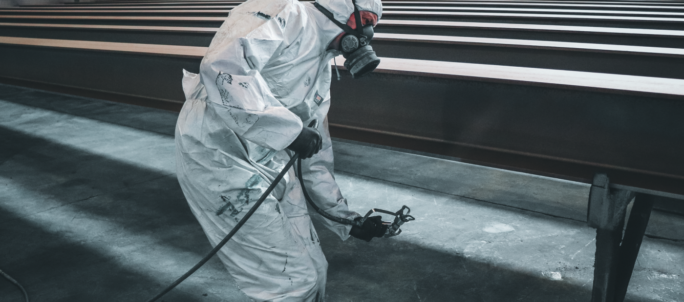 Worker in protective gear spray painting metal beams inside a factory setting.