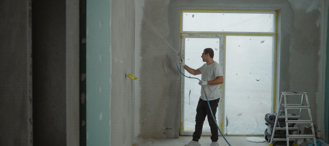 A man sprays paint on a wall while renovating an indoor room with professional equipment.